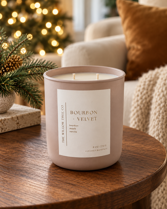 A large scented candle in a matte taupe jar sits on a wooden table in a cozy living room setting with a blurred Christmas tree and couch in the background. The label reads 'THE WILLOW TREE CO.