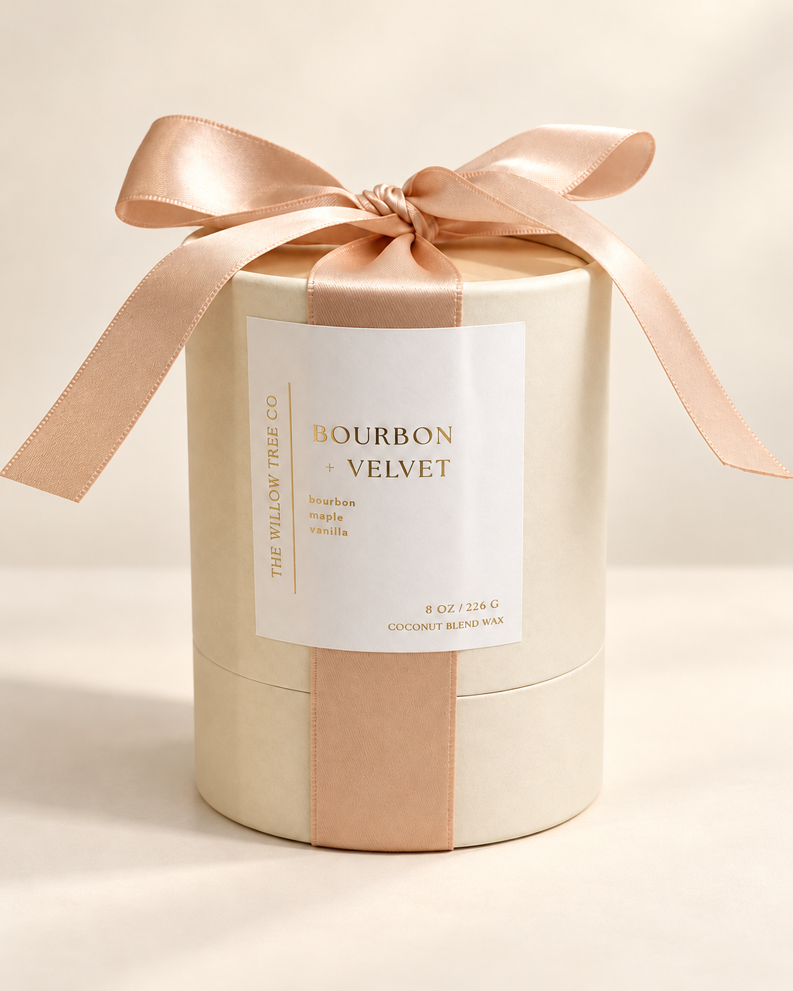 A cylindrical candle box wrapped in cream paper with a pale champagne colored ribbon and bow, featuring a white label that reads 'Bourbon + Velvet' and 'The Willow Tree Co.' on a warm cream background.