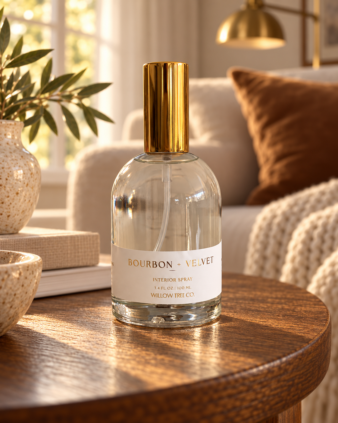 A clear glass perfume bottle with a shiny gold cap and a white label reading 'Bourbon Velvet' and 'Willow Tree Co.' sits on a round wooden table in a warm, sunlit living room setting.