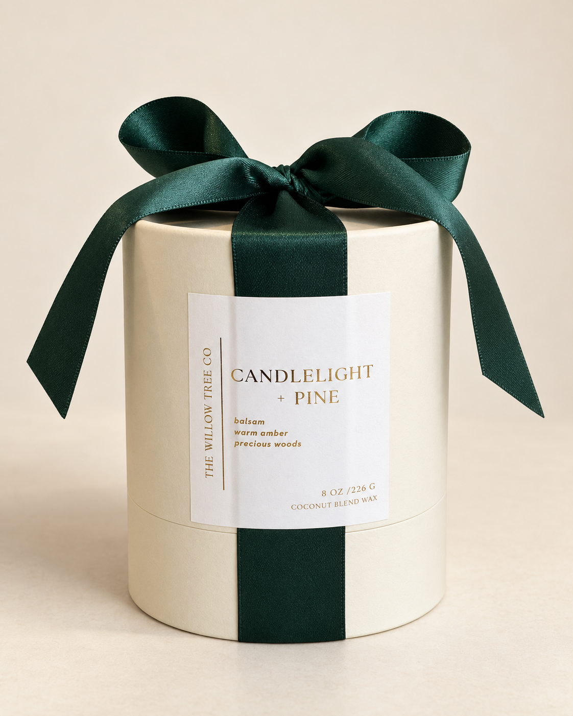A cylindrical candle box in warm cream with a white label and dark green ribbon, tied with a large bow. The label reads 'CANDLELIGHT + PINE' and shows 'The Willow Tree Co.