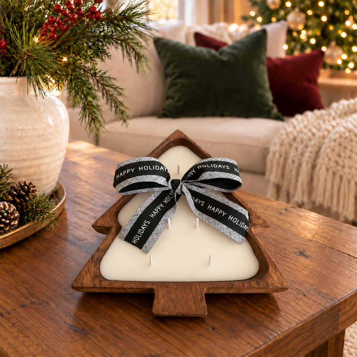 A Christmas tree-shaped candle in a wooden dough bowl sits on a wooden table in a cozy living room setting. The candle wax is off-white, and it is tied with a dark green and white ribbon that reads 'HAPPY HOLIDAYS'.