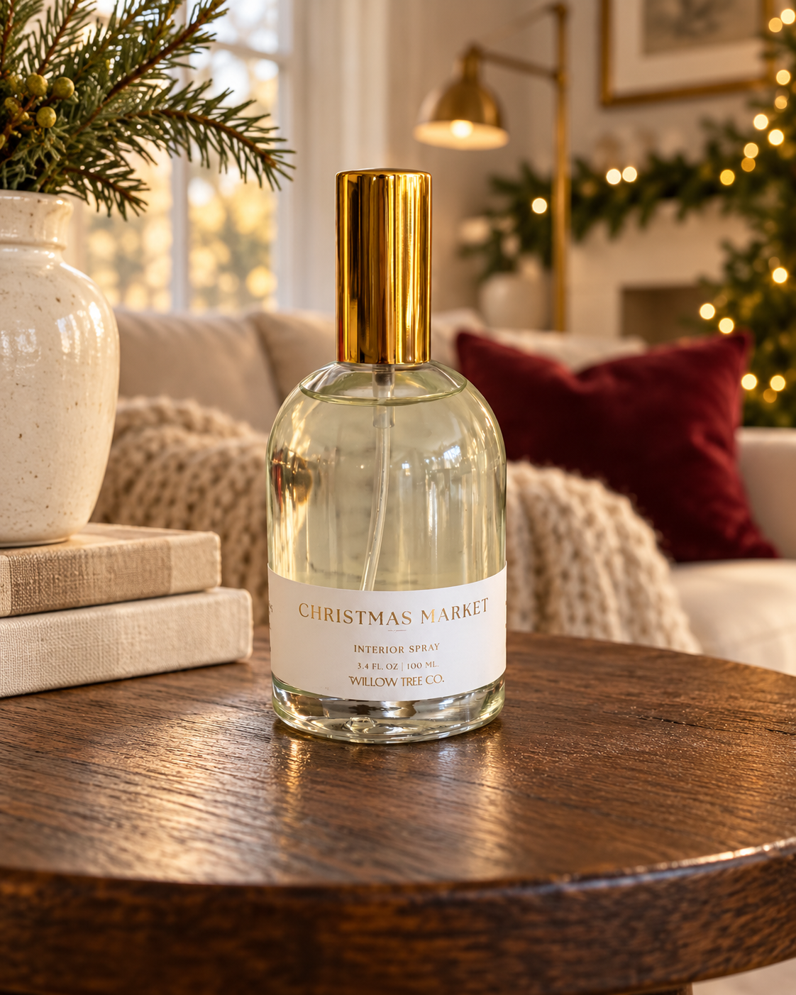 A clear glass bottle of Willow Tree Co. 'Christmas Market' interior spray with a gold cap and white label, set on a wooden table in a cozy, warmly lit living room with holiday decorations.