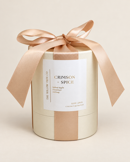 A cylindrical candle box wrapped in a champagne-gold satin ribbon tied in a bow at the top, with a white label reading 'CRIMSON + SPICE' and 'THE WILLOW TREE CO.' on a warm beige background.