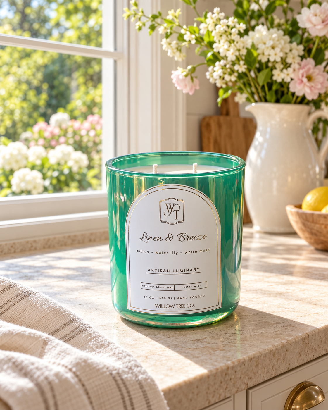 A green glass jar candle labeled 'Linen & Breeze' by Willow Tree Co., placed on a kitchen counter near a window with flowers and a vase in the background.