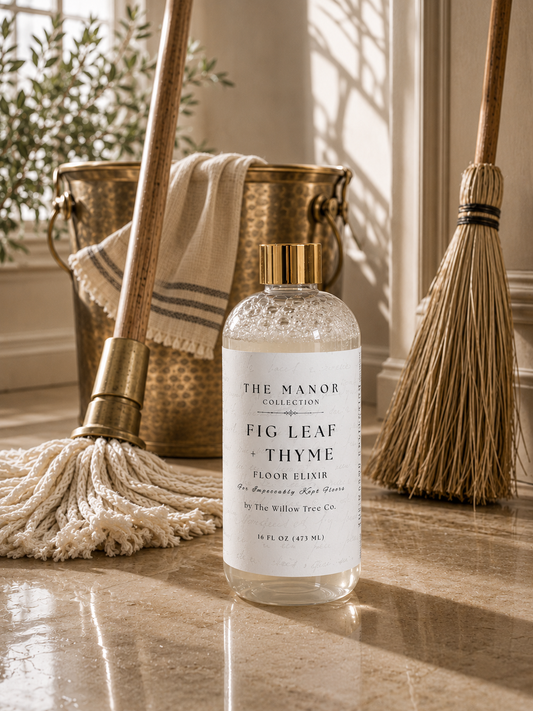 A bottle of Fig Leaf + Thyme Floor Cleaner by The Manor Collections stands on a polished stone floor, styled with a mop, broom, and a brass bucket with a towel in a sunlit interior.