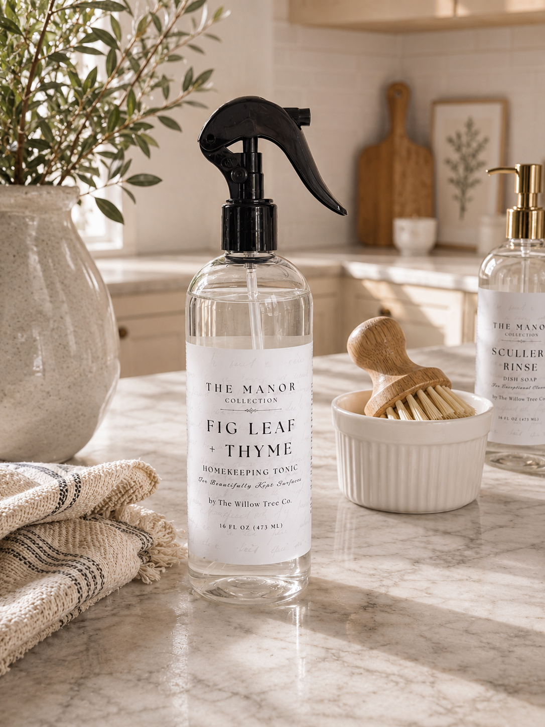 A clear spray bottle of The Manor Collection Fig Leaf + Thyme homreeping tonic with a black sprayer, set on a marble kitchen counter with a wooden brush and another bottle in the background.