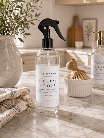 A clear spray bottle of The Manor Collection Fig Leaf + Thyme homreeping tonic with a black sprayer, set on a marble kitchen counter with a wooden brush and another bottle in the background.