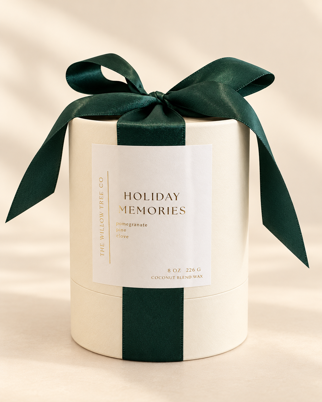 A cylindrical cream-colored candle tin wrapped with a dark green satin ribbon and bow, featuring a white label that reads 'HOLIDAY MEMORIES' by The Willow Tree Co.