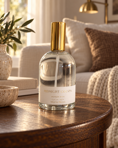 A clear glass bottle of Midnight Cocoa interior spray by Willow Tree Co sits on a round wooden table in a cozy living room setting with a sofa, throw pillow, and plant in the background.