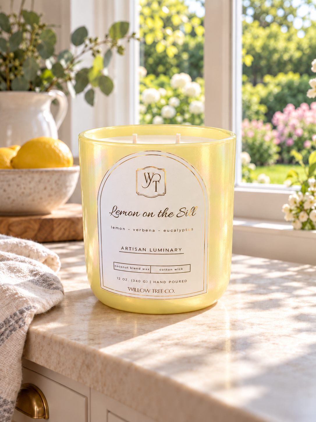 A yellow scented candle in a clear glass jar sits on a marble countertop in a bright kitchen or bathroom setting, with a window showing a green garden outside. The label reads 'Lemon on the Sill' by Willow Tree Co.
