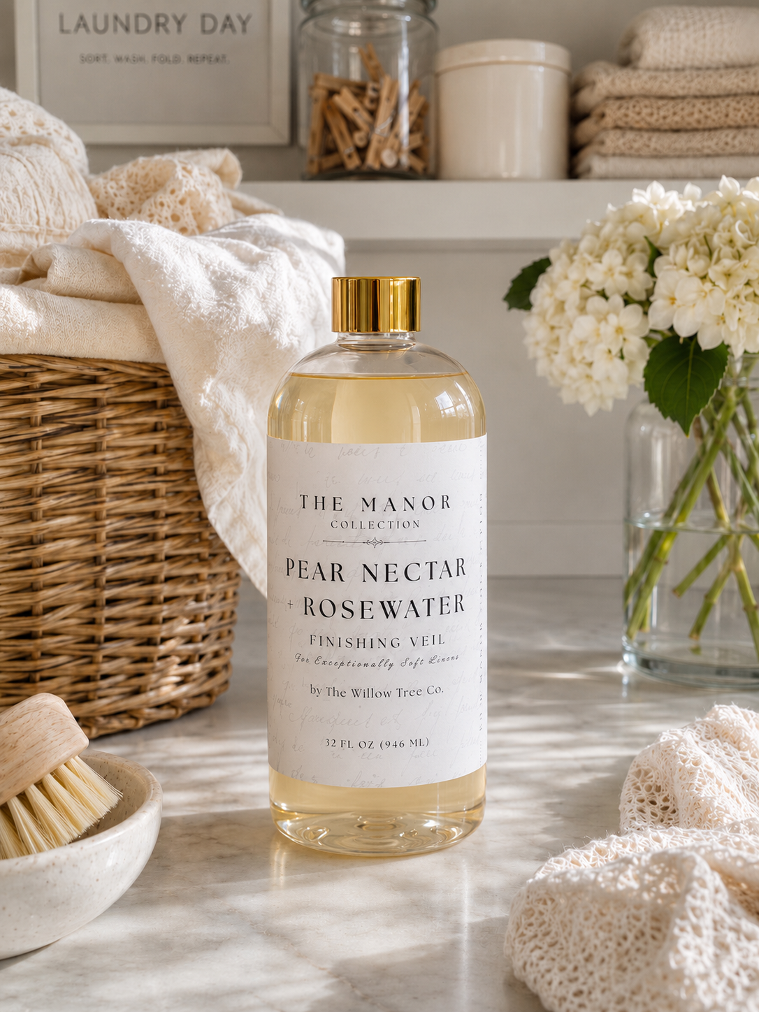 A bottle of The Manor Collection Pear Nectar Rosewater Finishing Alcohol stands on a marble countertop in a bright, styled laundry room setting with folded towels, a wicker basket, and white flowers.