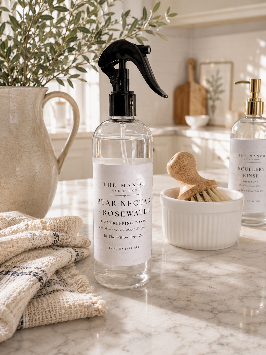 A clear spray bottle of The Manor Conservatory Pear Nectar Rosewater with a black sprayer sits on a marble countertop in a bright kitchen setting, alongside a folded beige cloth, a wooden brush in a white bowl, and another bottle in the background.