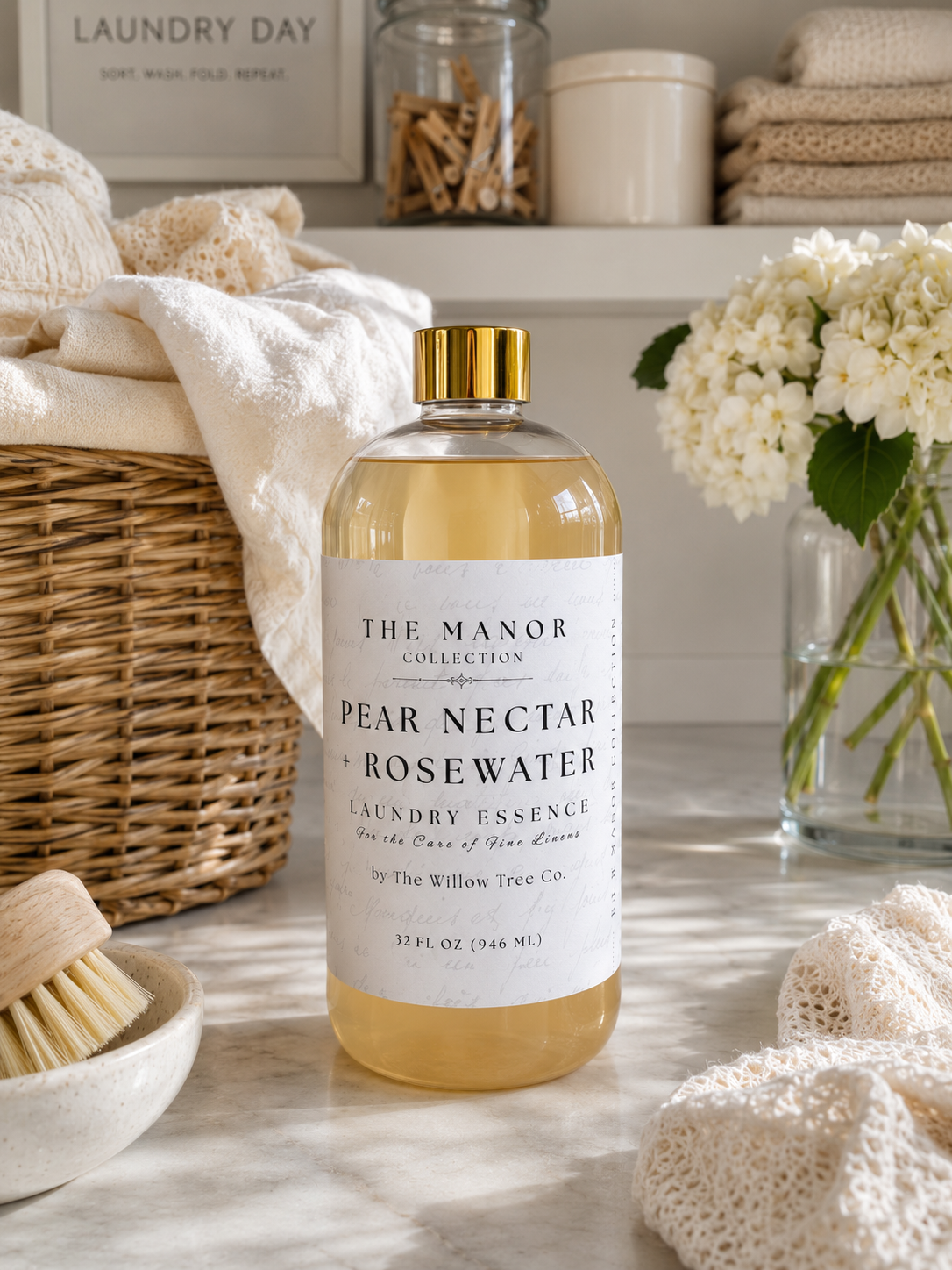 A bottle of The Manor Collection Pear Nectar + Rosewater Laundry Essence stands on a marble surface in a styled laundry room setting with towels, a wicker basket, flowers, and jars in the background.