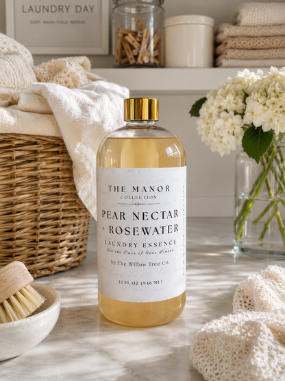 A bottle of The Manor Collection Pear Nectar + Rosewater Laundry Essence stands on a marble surface in a styled laundry room setting with towels, a wicker basket, flowers, and jars in the background.