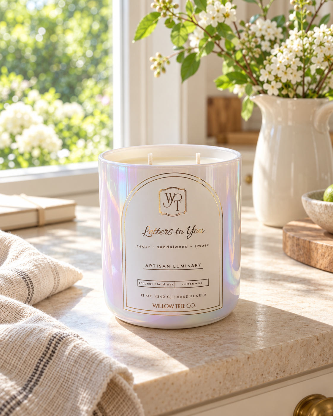 A lit scented candle in an iridescent white glass jar sits on a kitchen counter near a window, with a striped cloth nearby and flowers in a vase in the background. The label reads 'Letters to You' by Willow Tree Co.