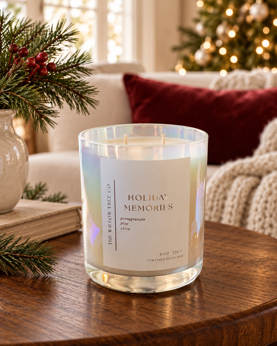 A holiday-themed scented candle in a clear glass jar with an iridescent finish, sitting on a wooden table in a cozy living room setting. The label reads 'HOLIDAY MEMORIES' and 'THE WILLOW TREE CO'.