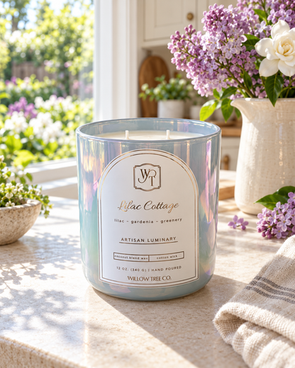 An iridescent blue-green scented candle in a glass jar sits on a sunlit kitchen counter, with a white label reading Lilac Cottage and Willow Tree Co. Purple flowers and a white pitcher are visible in the background near a window.