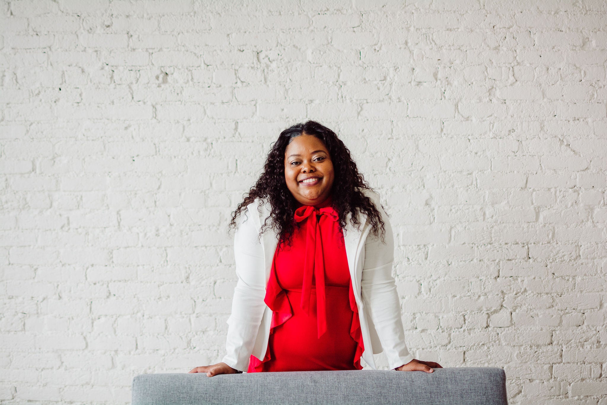 A young african american woman wearing a red dress and white blazer smiling while standing behing a medium gray sofa