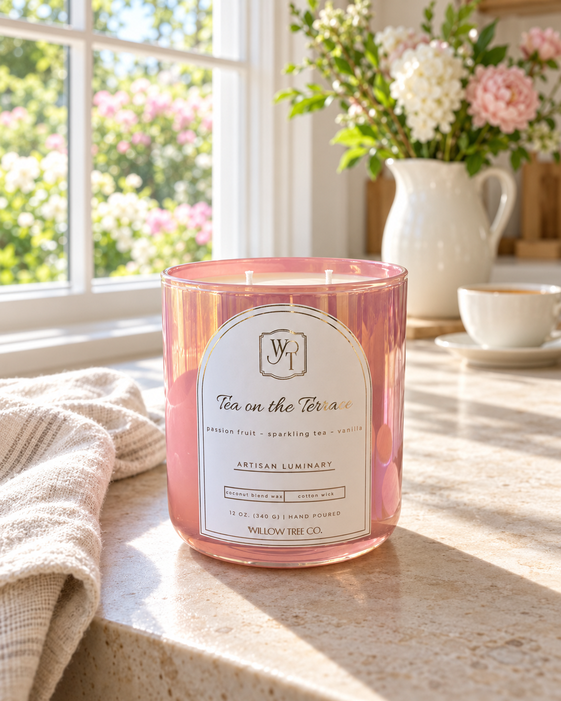 A pink iridescent glass jar candle labeled 'Tea on the Terrace' by Willow Tree Co., styled on a sunlit kitchen counter with a vase of flowers and a cup in the background.