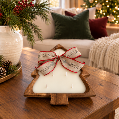 A Christmas tree-shaped candle in a wooden dough bowl sits on a wooden table in a cozy living room setting. The candle is white with a decorative red and beige ribbon bow tied around the top, and the background shows a decorated Christmas tree, pillows, and a sofa.