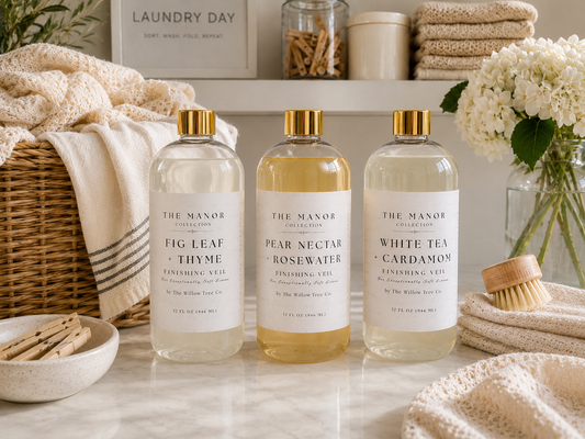 Three bottles of The Manor Collection body wash lined up on a marble countertop with a cozy, rustic bathroom setting featuring towels, a wicker basket, and white flowers.