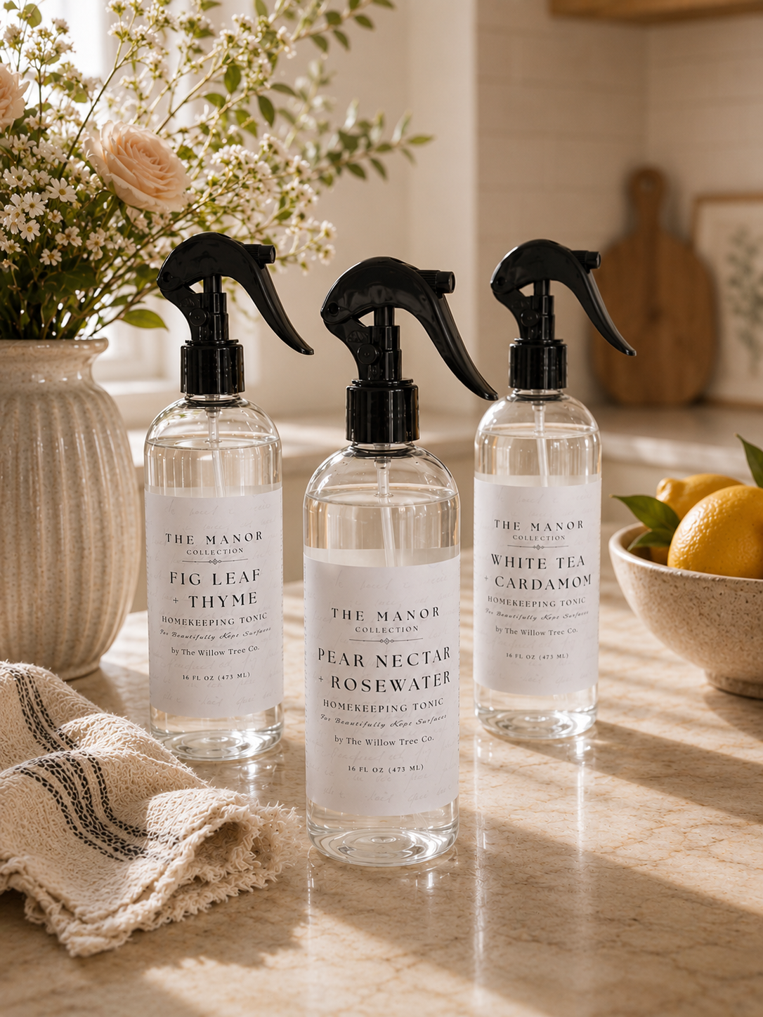 Three clear spray bottles of The Manor Botanicals room spray with black triggers and white labels, arranged on a marble countertop with flowers, a towel, and lemons in a warm kitchen setting.