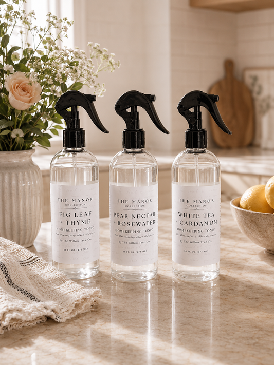 Three clear spray bottles of The Manor London room spray with black triggers and white labels, arranged on a marble countertop in a warm kitchen setting with flowers, a towel, and lemons.
