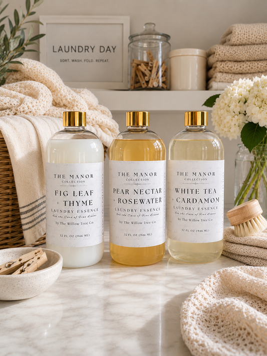 Three bottles of The Manor Collection laundry essences (Fig Leaf Thyme, Pear Nectar Rosewater, and White Tea Cardamom) lined up on a marble countertop with cozy laundry-themed props like a basket, folded towels, and a soap dish.