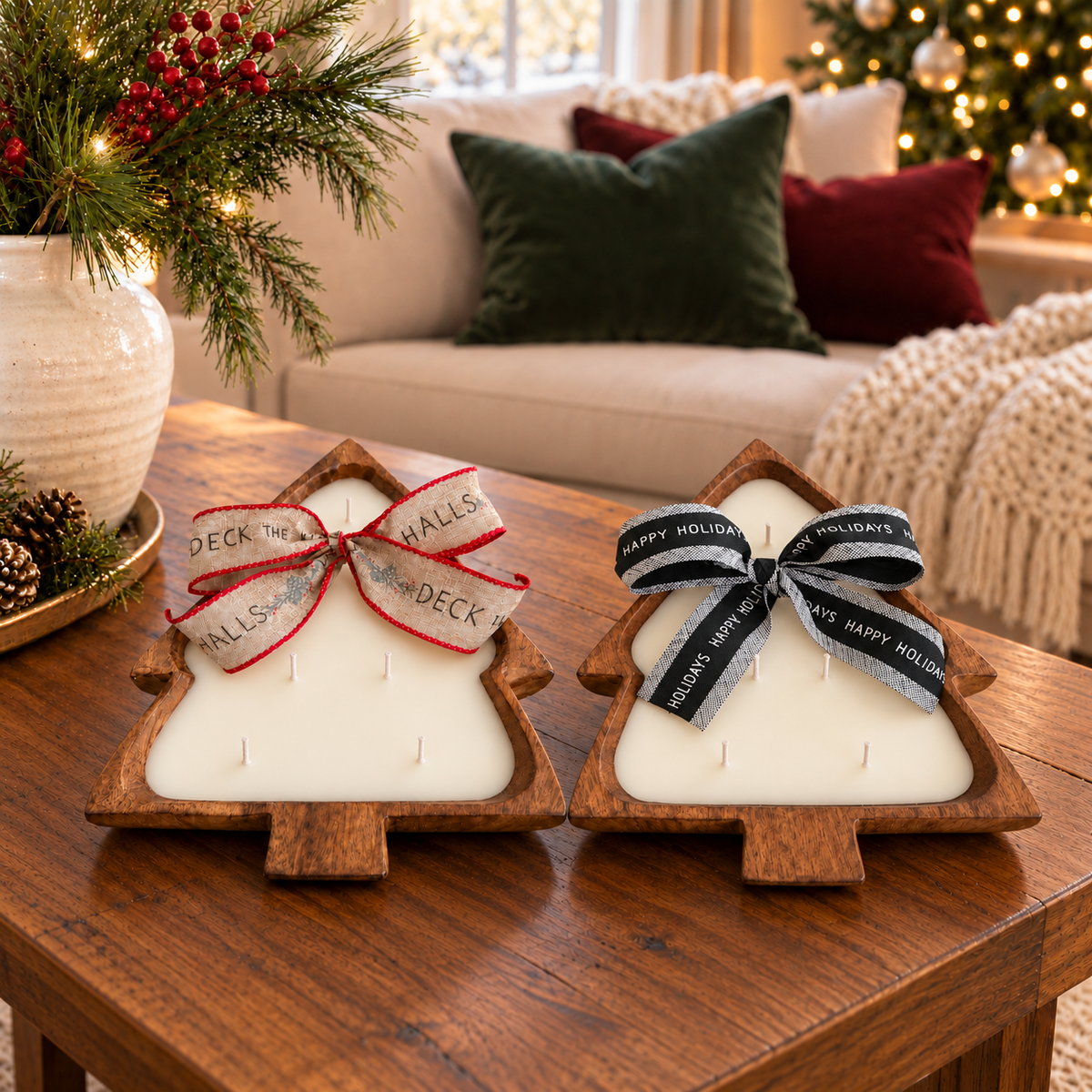 Two Christmas tree-shaped wooden dough bowl candles with five cotton wicks each and festive ribbon bows on a wooden coffee table, beside an ivory vase of evergreen branches.