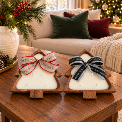 Two Christmas tree-shaped wooden dough bowl candles with five cotton wicks each and festive ribbon bows on a wooden coffee table, beside an ivory vase of evergreen branches.