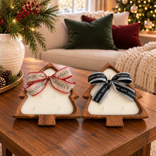 Two Christmas tree-shaped wooden dough bowl candles with five cotton wicks each and festive ribbon bows on a wooden coffee table, beside an ivory vase of evergreen branches.
