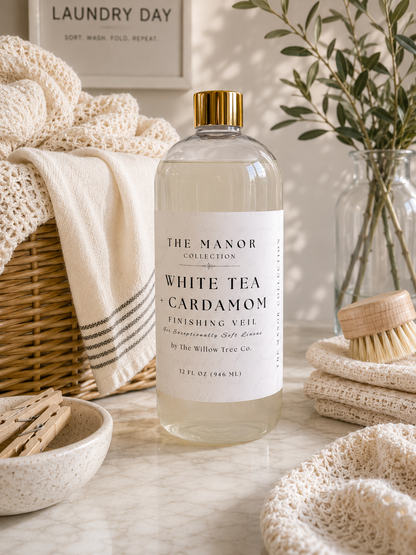 A clear bottle of The Manor Collection White Tea + Cardamom finishing veil hair product with a gold cap, styled on a marble surface with a wicker basket, cream knit textiles, a brush, and dried greenery in the background.