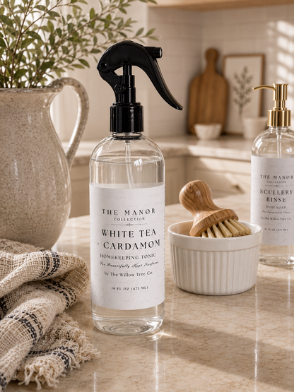 A clear glass spray bottle of The Manor Collection White Tea + Cardamom homekeeping tonic with a black trigger sprayer, styled on a beige marble countertop with a burlap towel, a wooden brush, and another bottle in the background.