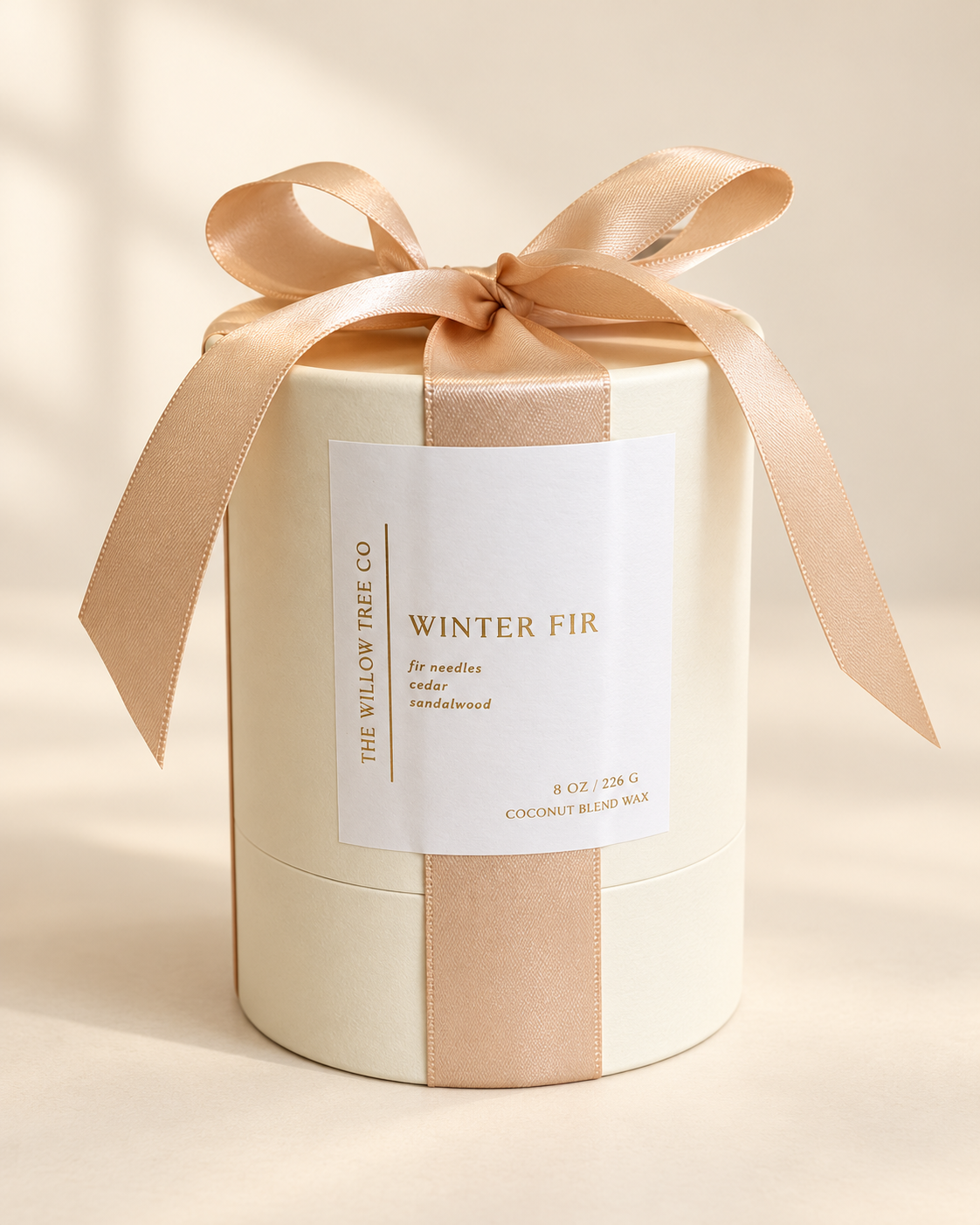 A cylindrical candle box wrapped in a cream-colored paper sleeve with a beige satin ribbon and bow, featuring a white label that reads 'WINTER FIR' and 'THE WILLOW TREE CO.'.