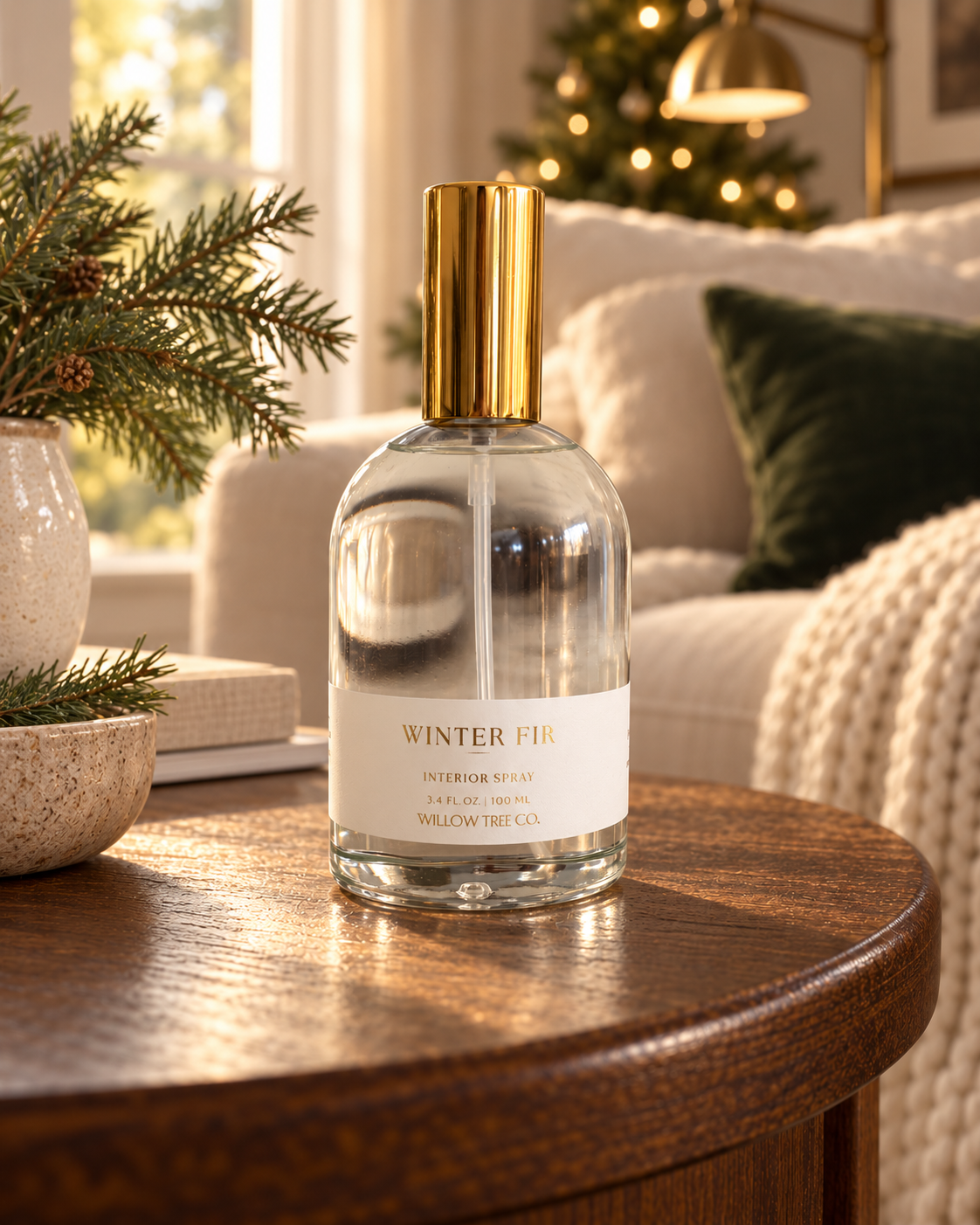 A clear glass bottle of Willow Tree Co. Winter Fir Interior Spray with a gold cap and white label, set on a wooden table in a cozy, softly lit living room with a Christmas tree in the background.