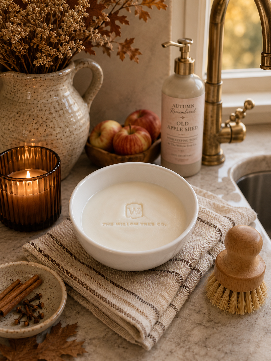 A white ceramic candle bowl with a wooden wick sits on a striped beige towel on a kitchen counter, surrounded by fall-themed props including a lit candle, apples, a pitcher of dried flowers, and a soap dispenser labeled Autumn Brewed with Old Apple Spice.