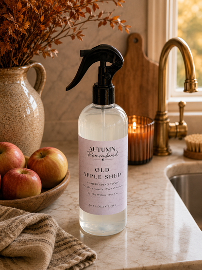 A clear spray bottle of Old Apple Shed home fragrance with a black trigger sprayer and a pale pink label, standing on a marble kitchen counter near a brass faucet, with apples, a candle, and dried flowers in the background.
