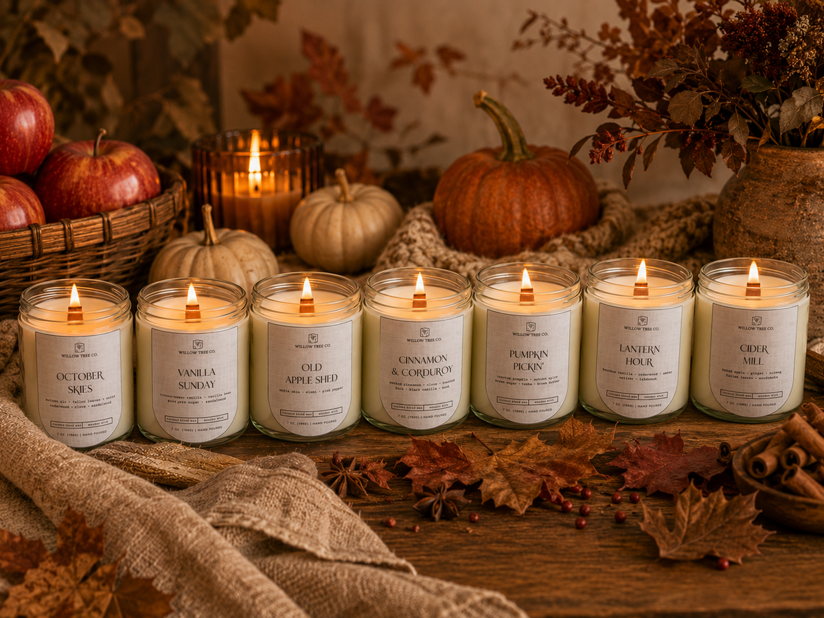 Seven lit scented candles in clear glass jars are arranged on a rustic wooden surface with autumn props including pumpkins, apples, leaves, and burlap. The labels show different scent names such as October Sage, Vanilla Sunday, Elder Apple Pie, Cinnamon & Cloves, Pumpkin Pie, Landon Rose, and Crux Me.