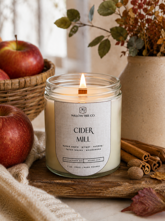 A lit candle in a clear glass jar labeled 'CIDER MILL' by Yellow Tree Co., styled on a wooden board with apples, cinnamon sticks, dried leaves, and a neutral fabric in a warm autumn setting.