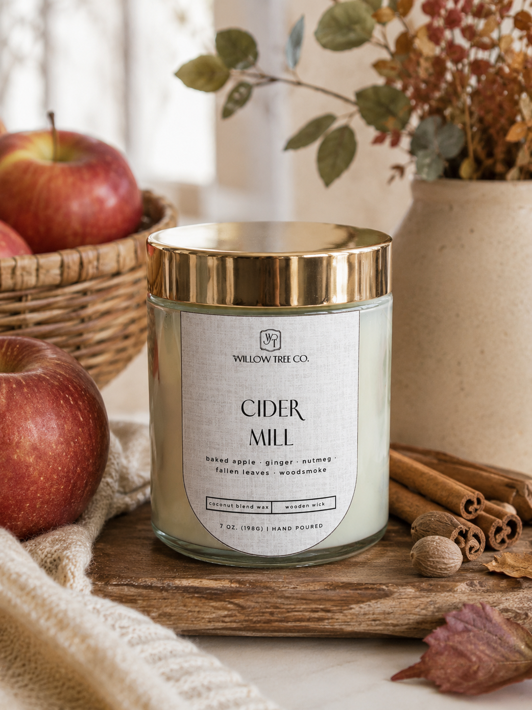 A candle in a clear glass jar with a gold lid and a white label reading 'CIDER MILL' by Willow Tree Co., styled on a wooden board with apples, cinnamon sticks, and dried flowers in the background.
