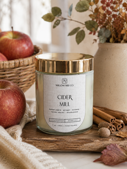 A candle in a clear glass jar with a gold lid and a white label reading 'CIDER MILL' by Willow Tree Co., styled on a wooden board with apples, cinnamon sticks, and dried flowers in the background.