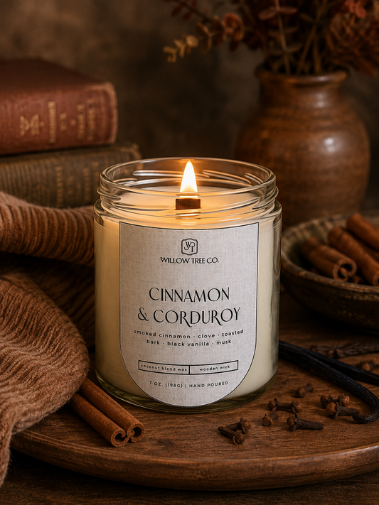 A lit candle in a clear glass jar with a cream label reading 'WILLOW TREE CO. CINNAMON & CORDUROY,' styled on a wooden surface with cinnamon sticks, books, and a warm rustic background.