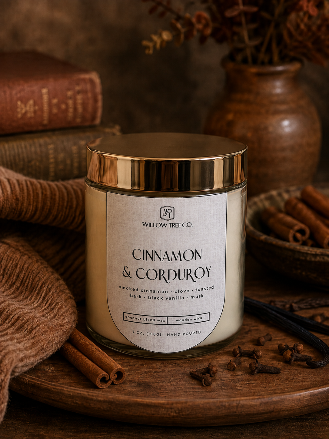 A jar candle labeled 'Cinnamon & Corduroy' by Willow Tree Co., styled on a wooden tray with cinnamon sticks, a knit fabric, books, and a ceramic vase in a warm rustic setting.