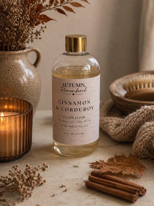 A bottle of Autumn Remembered Cinnamon & Corduroy floor elixir by The Willow Tree Co. styled on a rustic tabletop with a lit candle, dried flowers, cinnamon sticks, and a knitted blanket.