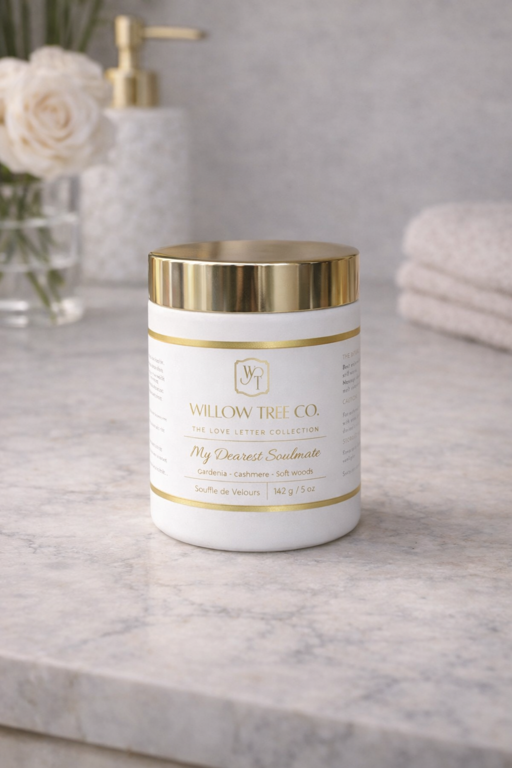Candle with gold lid and 'Willow Tree Co.' label on a marble surface with flowers and towels in the background.