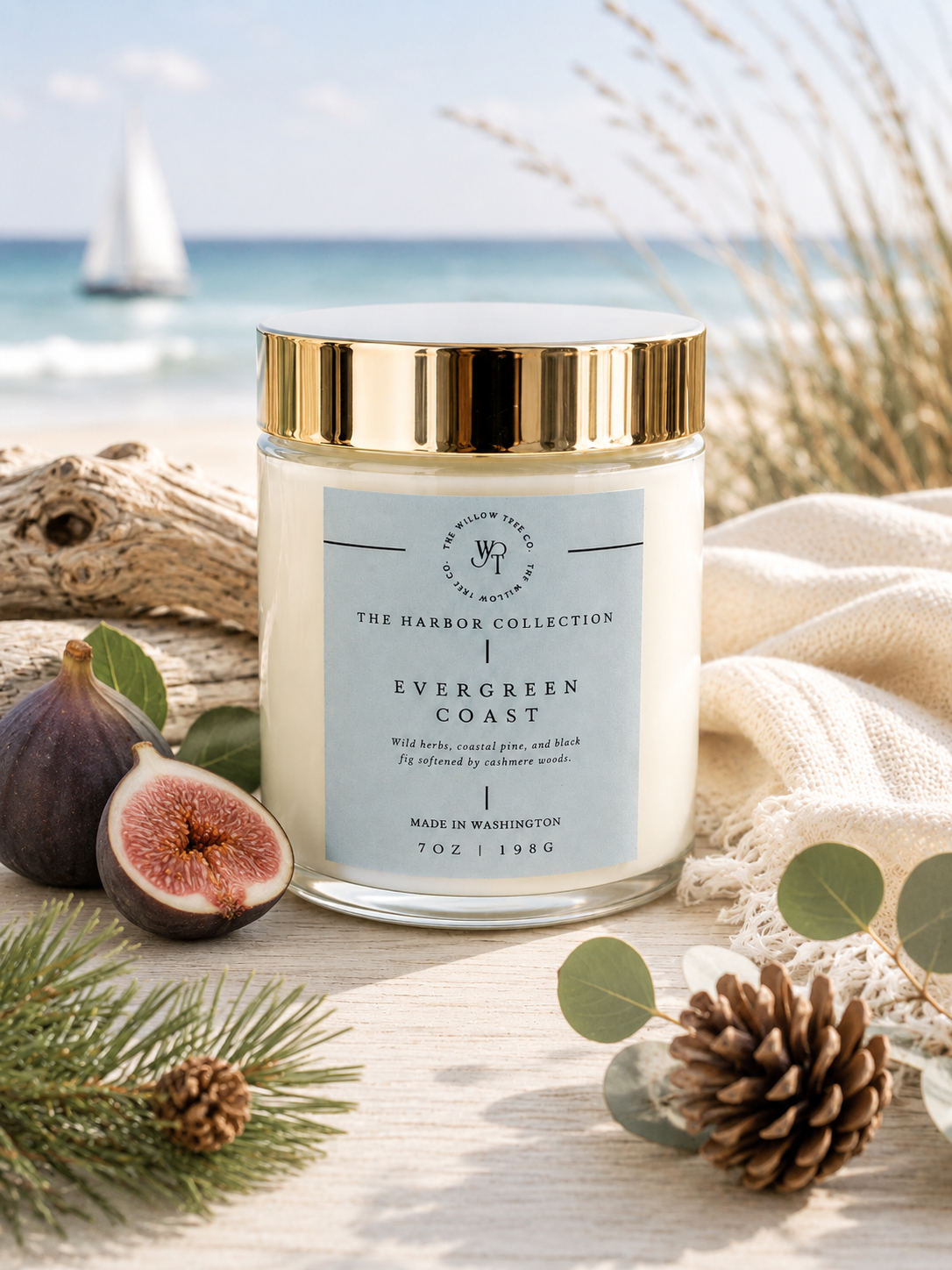 Candle labeled 'Evergreen Coast' from The Harbor Collection on a beach setting with ocean and sailboat in the background.