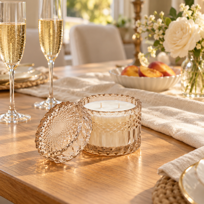 A candle in a textured, faceted golden glass container sits on a wooden dining table with its matching lid beside it. The scene is styled with champagne flutes, a folded cream towel, flowers, and fruit in a warm, bright interior.