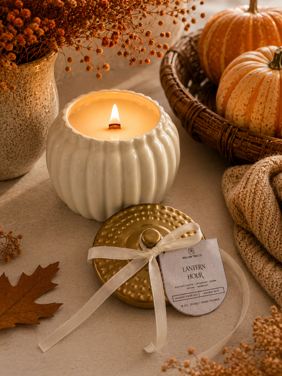 A lit candle in a ribbed white pumpkin-shaped holder sits beside its gold lid and a product tag reading 'Lantern Hour' on a beige fabric surface. Pumpkins, dried flowers, a wicker basket, and a knitted beige scarf create a warm autumn styled scene.