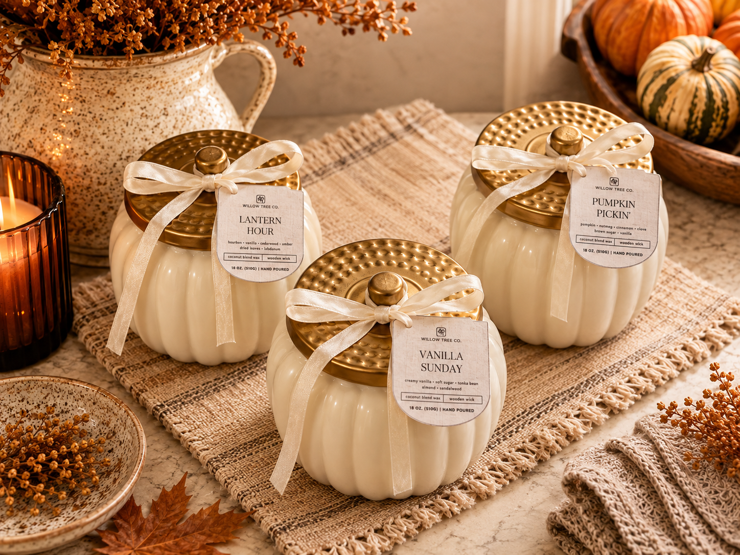 Three decorative candle jars shaped like pumpkins with gold lids and cream ribbed bodies, tied with white ribbons and labeled 'Lantern Hour,' 'Vanilla Sunday,' and 'Pumpkin Pickin'. They are styled on a woven runner with autumn props including pumpkins, a candle, and dried flowers.