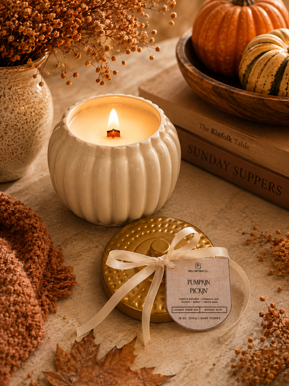 A lit cream-colored ribbed candle in a pumpkin-shaped holder sits on a tabletop beside a gold tin lid tied with a cream ribbon and a tag reading 'Pumpkin Pickin.' The scene is styled with autumn props including dried flowers, a knit blanket, books, and small pumpkins.